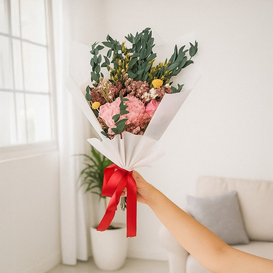 Lifestyle image showing preserved dried flower bouquet wrapped in white paper with red ribbon held by hand in bright modern living room setting.