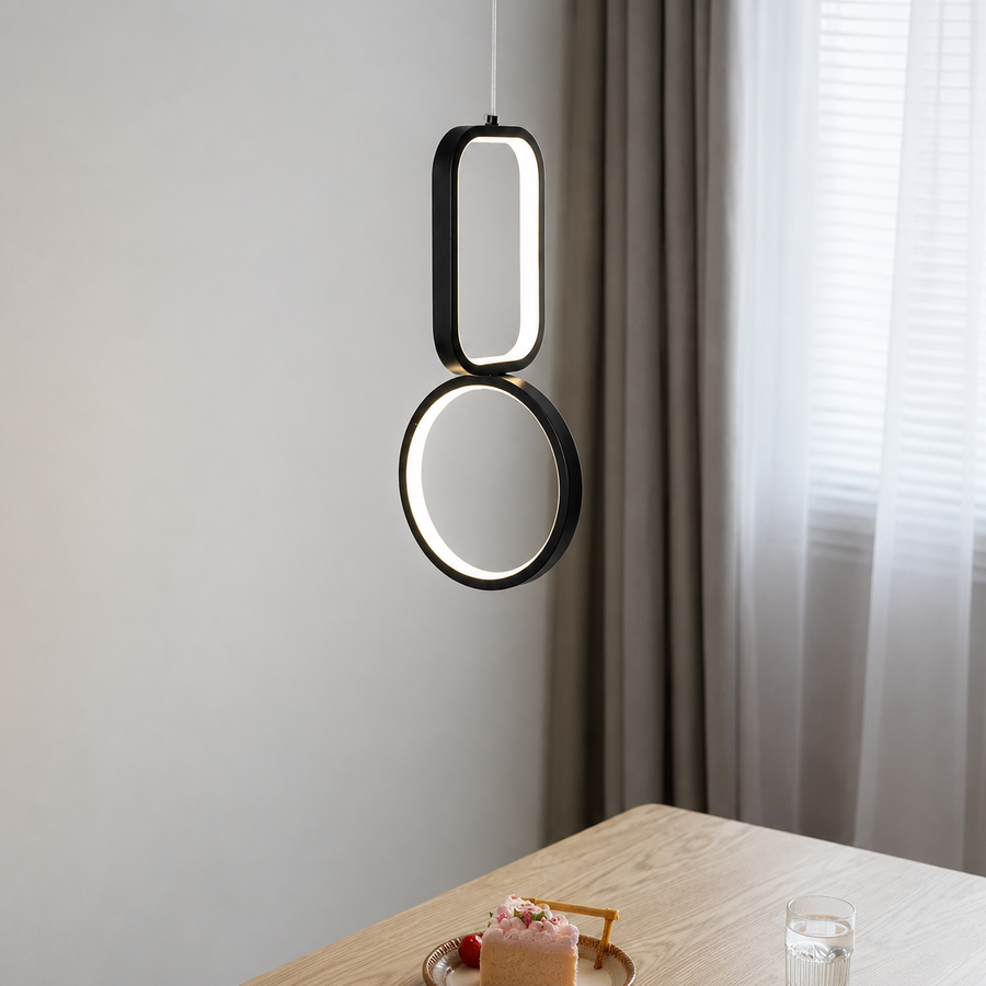 A modern black pendant light with two stacked oval and circular illuminated sections hanging over a wooden dining table in a softly lit room with curtains.