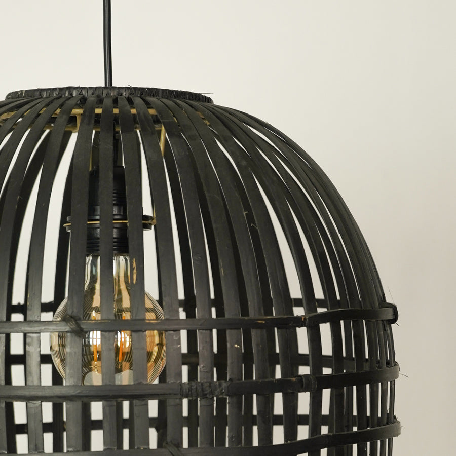 A close-up of a black woven rattan or wicker birdcage-style pendant light with a visible warm-toned Edison bulb inside, hanging against a plain light background.