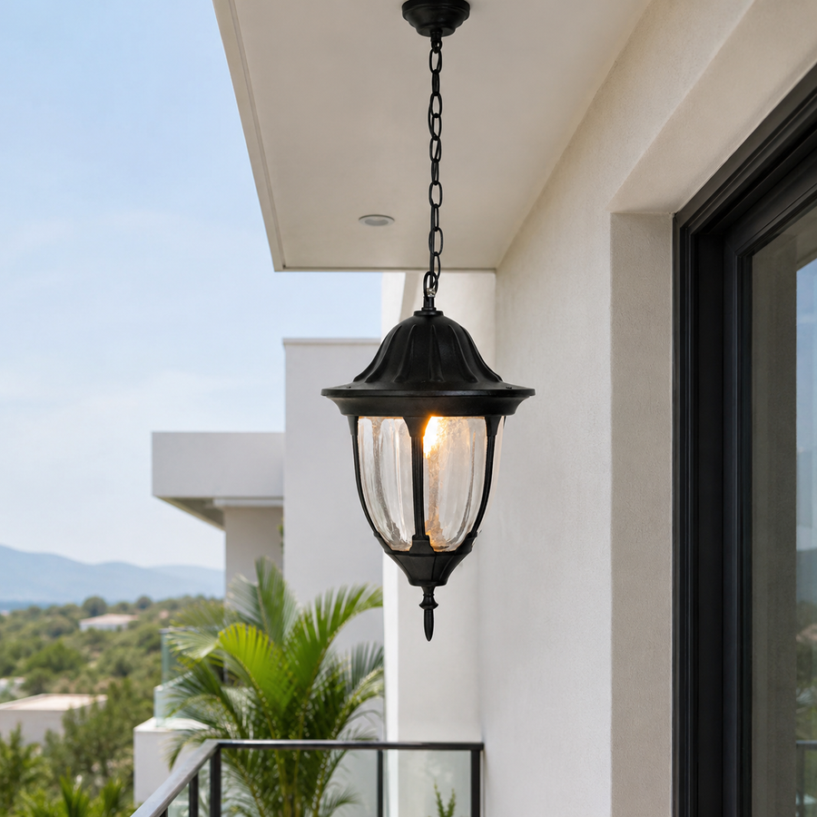 A black outdoor pendant light fixture hanging from a chain under a white building eave, with a warm glowing bulb inside clear glass panels. Palm leaves and distant hills are visible in the background.
