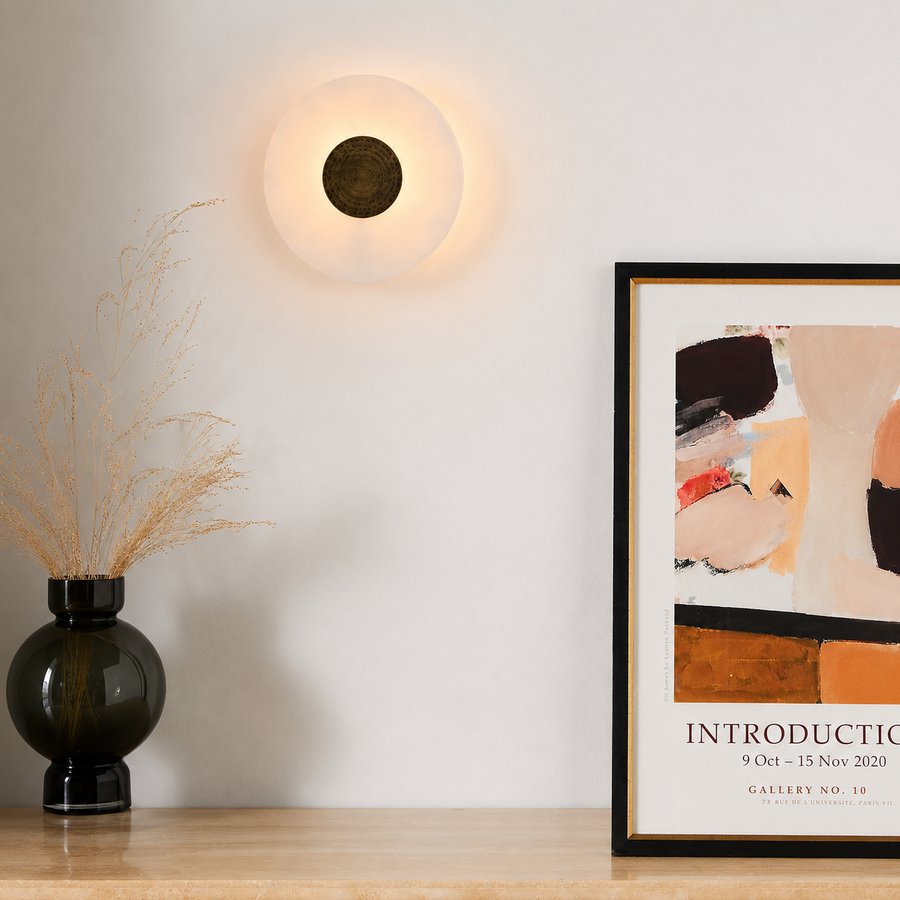 A modern wall-mounted circular light fixture glowing warm white on an off-white wall, shown above a wooden surface with a black vase holding dried grass and a framed abstract art print.