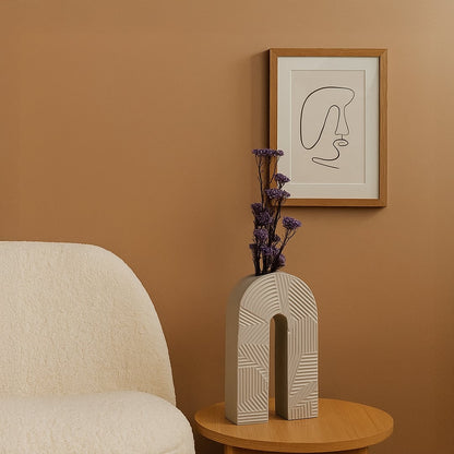 White ceramic arch vase with geometric texture holding purple flowers on a wood table next to a boucle chair for modern decor.