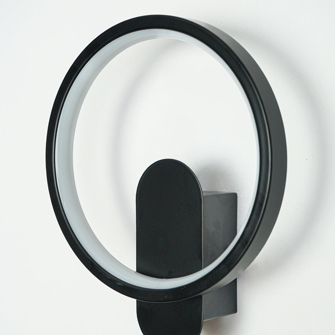 Close view of black metal wall sconce with circular LED ring and oval backplate on clean white wall background showcasing contemporary lighting design.