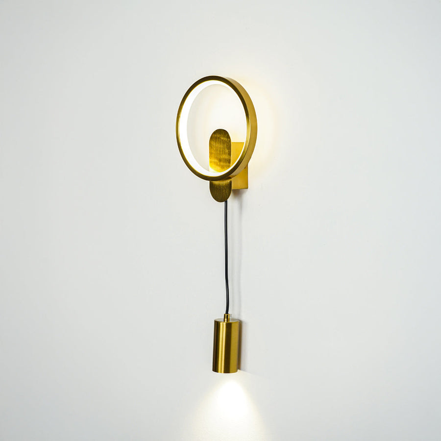 Minimal gold wall sconce with round frame and cylindrical spotlight shade on clean neutral wall background perfect for contemporary interior lighting.