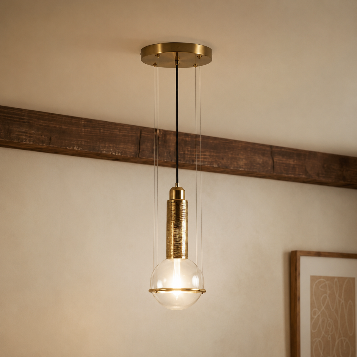 A modern pendant light fixture with a brushed brass circular ceiling canopy, a black suspension cord, a cylindrical brass shade, and a clear glass globe bulb, shown hanging in a room with a beige wall and a dark wood beam.
