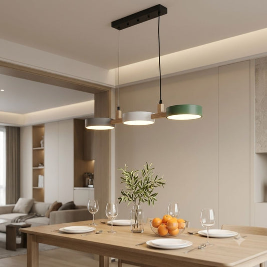 A modern three-light linear pendant chandelier with circular shades in black, white, and sage green, hanging over a wooden dining table set for a meal in a contemporary dining room.