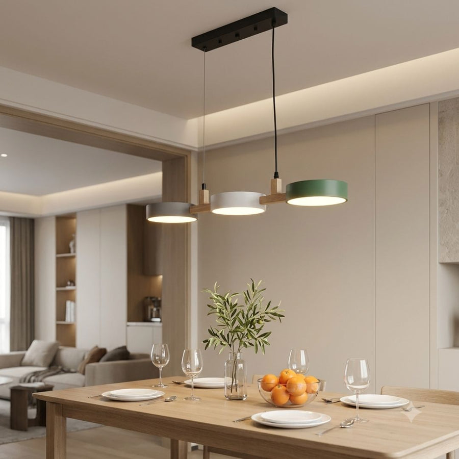 A modern three-light linear pendant chandelier with circular shades in black, white, and sage green, hanging over a wooden dining table set for a meal in a contemporary dining room.