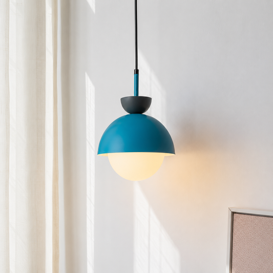 A modern pendant light with a matte teal dome shade, a black top cap, and a glowing warm white globe bulb, hanging against a white wall with soft sunlight and curtains.