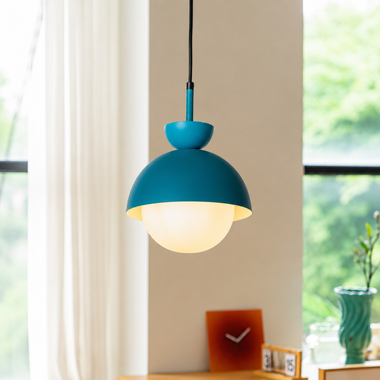 A modern pendant light with a matte teal-blue dome shade and a frosted white globe bulb, hanging in a bright room with a window, curtain, and small tabletop decor in the background.