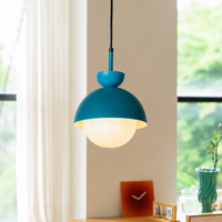 A modern pendant light with a matte teal-blue dome shade and a frosted white globe bulb, hanging in a bright room with a window, curtain, and small tabletop decor in the background.