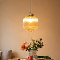 Contemporary gold brass pendant light with a glowing vertical ribbed amber glass shade hanging in a dining room above a floral arrangement.