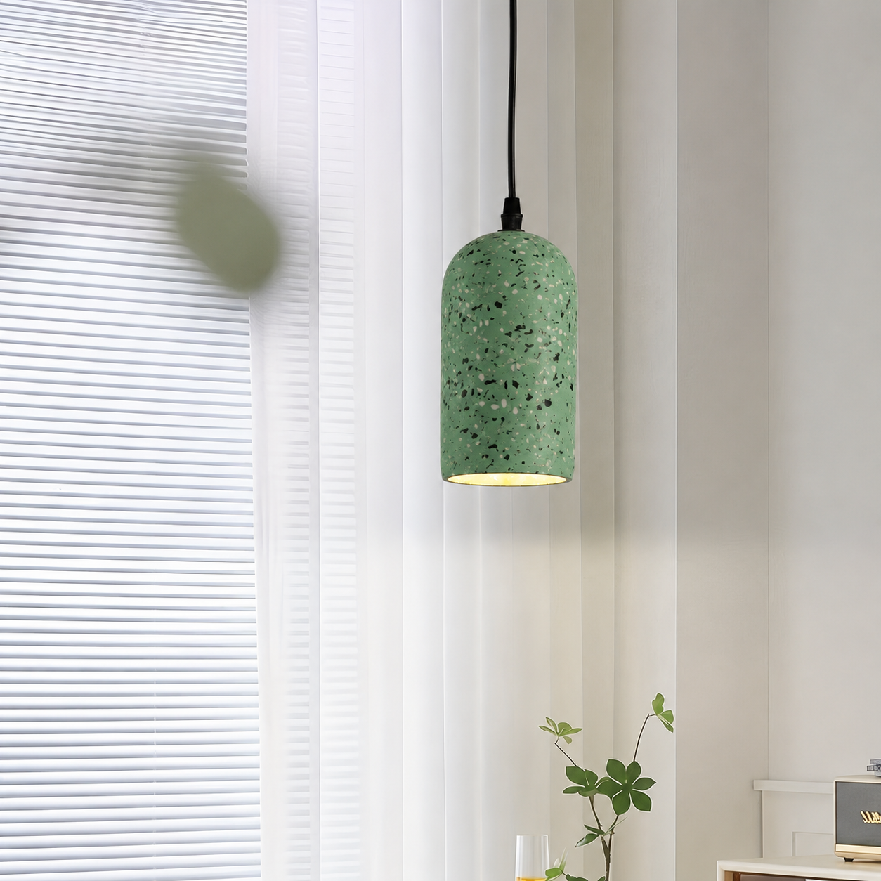 A green speckled pendant light with a terrazzo-style finish hangs in a bright room with white blinds and a small plant below.