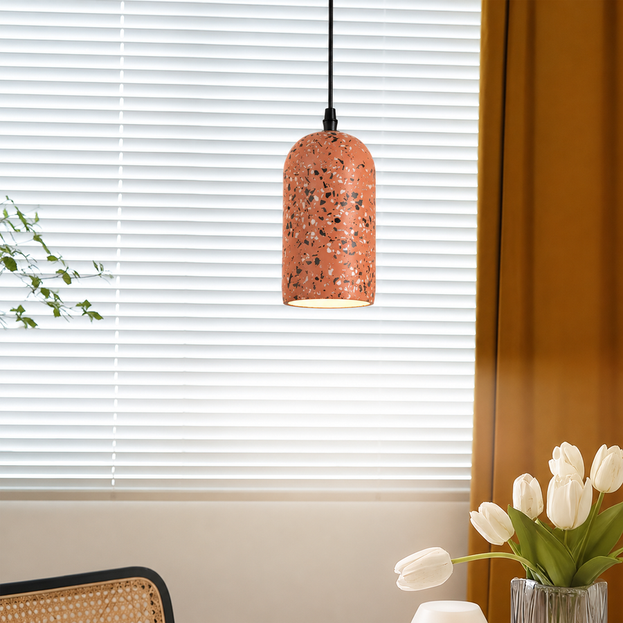 A terrazzo-patterned pendant light with a copper-orange shade hangs in front of white window blinds in a styled room setting with a vase of white tulips and a mustard curtain.