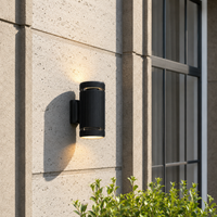 Modern black aluminum cylinder downlight wall sconce fixture on concrete exterior wall next to house glass window decor.