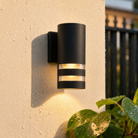 Modern black metal cylindrical grid downlight wall sconce fixture on cream stucco pillar for contemporary patio lighting.