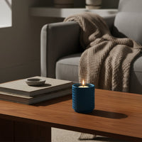 Blue textured candle burning on wooden coffee table with stacked books beside soft sofa and knit throw creating calm cozy living room decor.