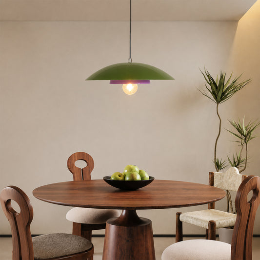 A green dome-shaped pendant light hangs above a round wooden dining table in a modern room with chairs, a bowl of green apples, and potted plants against a beige wall.