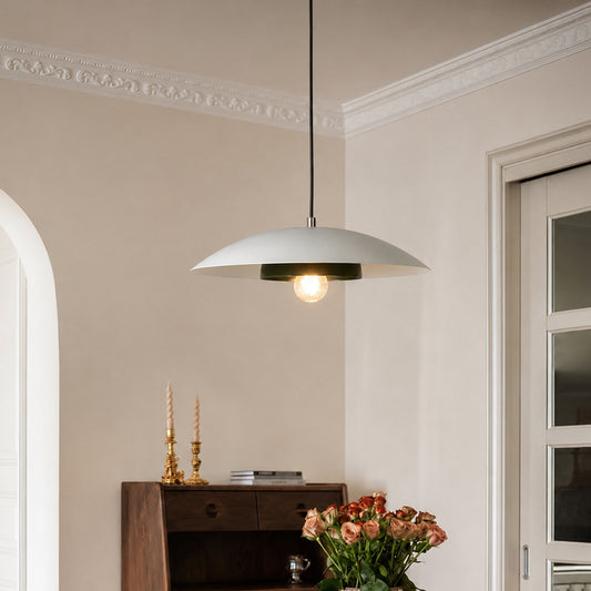 A modern pendant light with a wide, shallow white dome shade and a black cord, hanging in a styled room with beige walls, a wooden cabinet, and a vase of pink roses.