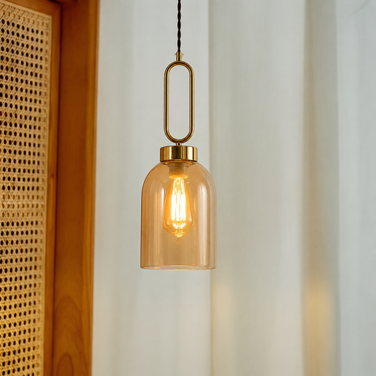 A modern pendant light with a clear amber-tinted glass bell shade and a brass-toned metal loop hanger, suspended in front of a softly lit interior background.
