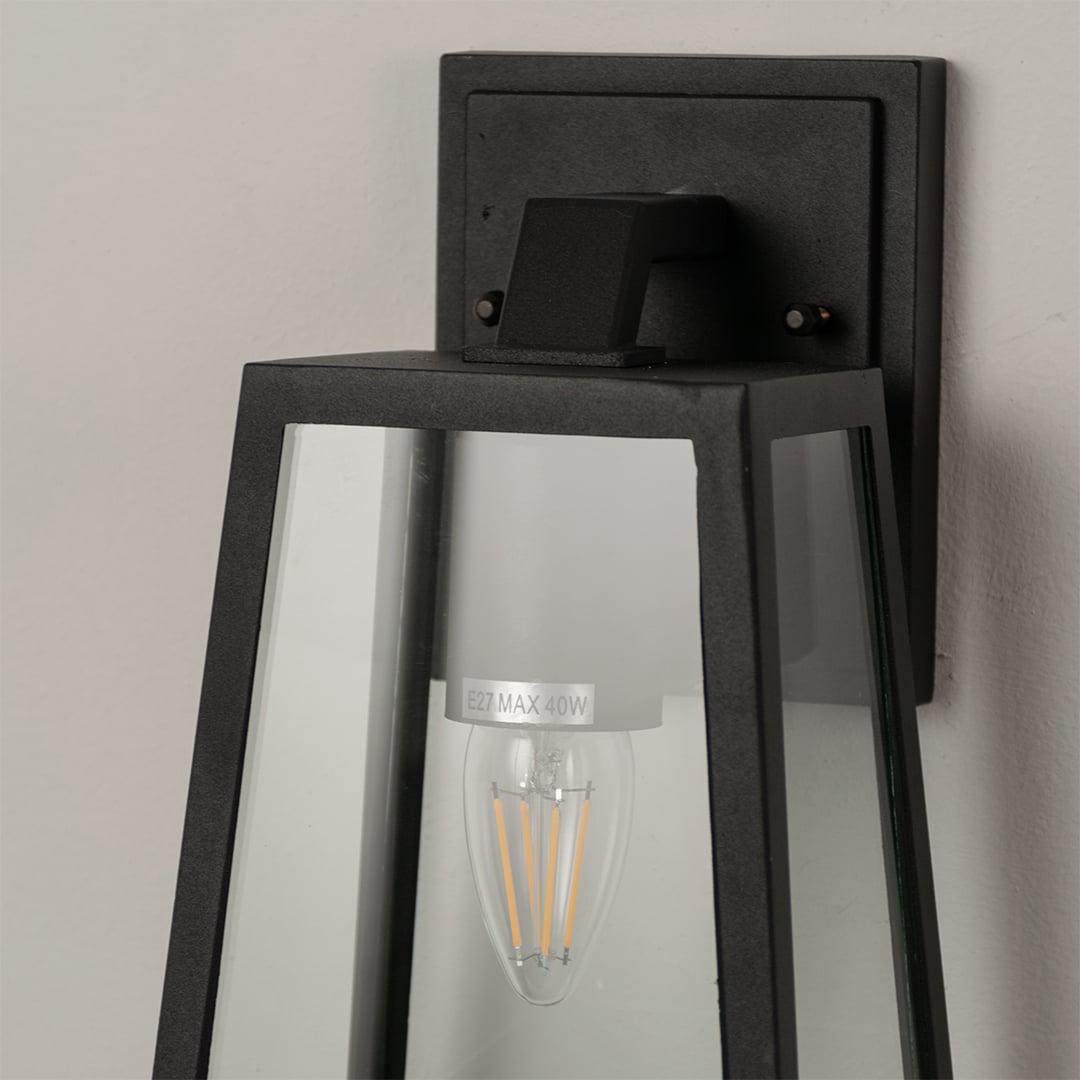 Closeup of black metal outdoor wall lantern with clear glass panels showcasing bulb detailing and modern geometric design.