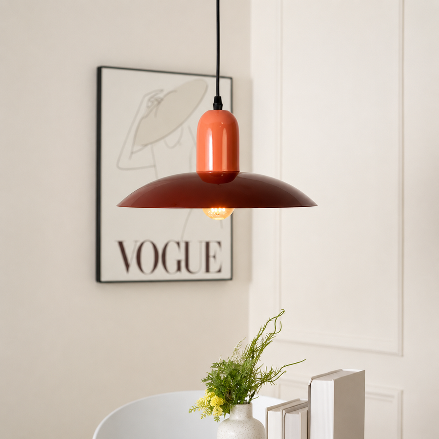 A modern pendant light with a glossy terracotta orange cylindrical shade and a wide matte dark red-brown metal dome shade, hanging in a styled interior setting with a framed VOGUE print on the wall.
