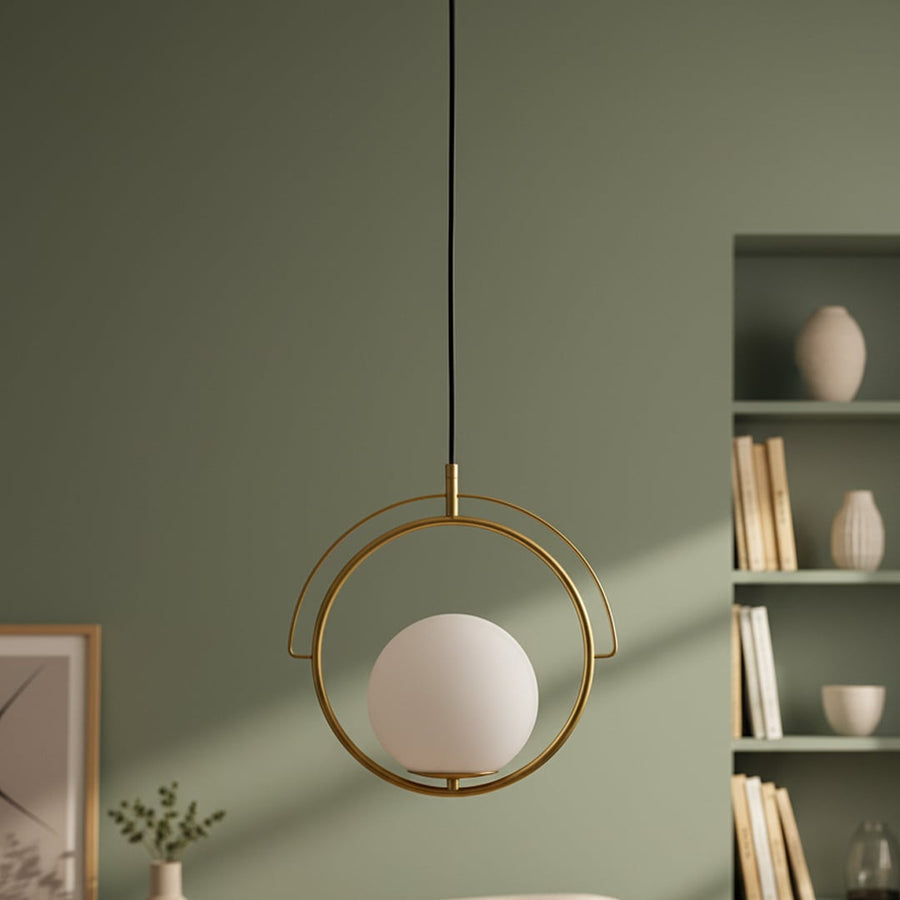 A decorative pendant light with a round white globe and gold circular frame hanging in a modern room with soft green walls and styled shelves.
