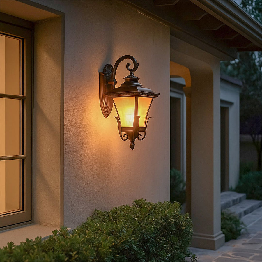 A classic outdoor wall lantern light glowing warmly at the entrance of a home with soft illumination cast on the wall and surrounding greenery during evening time.