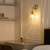 A warm brass wall sconce with a textured clear glass globe shade, mounted on a cream-colored wall in a bedroom setting.