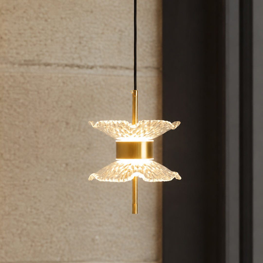 A modern pendant light with two textured, petal-like clear glass shades and a brushed gold metal center and rod, suspended by a black cord against a textured beige wall and dark frame.