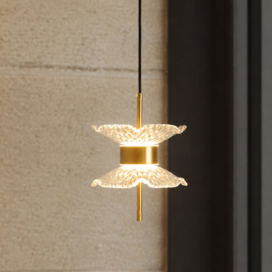 A modern pendant light with two textured, petal-like clear glass shades and a brushed gold metal center and rod, suspended by a black cord against a textured beige wall and dark frame.