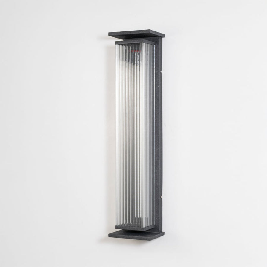 Black ribbed wall light with vertical glass shade displayed off on plain white background surface.