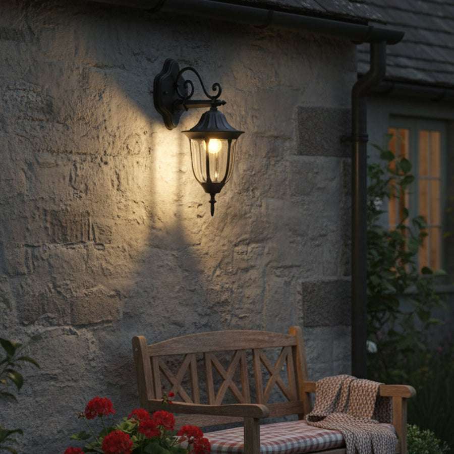 Outdoor wall light with black metal lantern design mounted on stone exterior wall casting warm light over garden seating area and evening home facade ambiance.