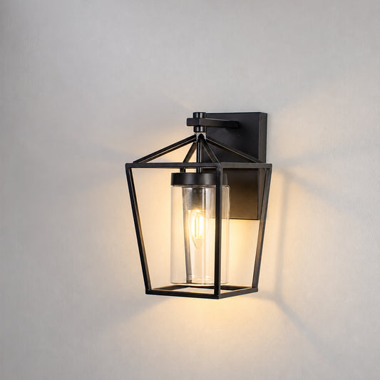 A black metal lantern-style wall sconce with a clear glass shade and visible warm Edison bulb, mounted on a light gray wall.
