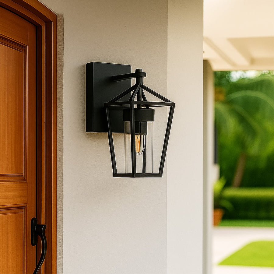 Outdoor wall light in black metal with clear glass mounted beside wooden door showing classic lantern design for exterior home lighting.