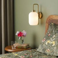 Gold metal wall light with white ribbed glass shade casting warm glow styled beside bed for cozy bedroom wall decor ambiance.