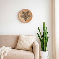 A round woven basket hangs on a white wall above a beige sofa with a knit pillow beside a tall green snake plant in a white planter.