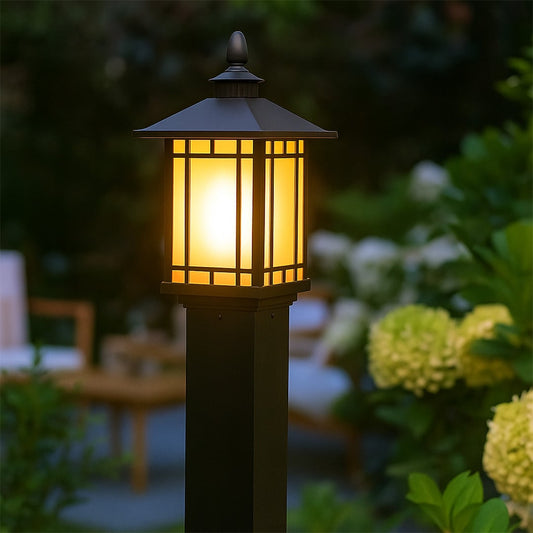 A modern outdoor garden scene featuring a tall black pillar lamp with the light on casting a warm glow across lush plants and seating areas creating a cozy evening atmosphere.