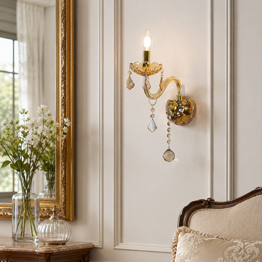 An ornate gold wall sconce with crystal hanging drops, mounted on a cream paneled wall in a styled interior next to a mirror and a vintage sofa.