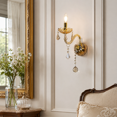 An ornate gold wall sconce with crystal hanging drops, mounted on a cream paneled wall in a styled interior next to a mirror and a vintage sofa.