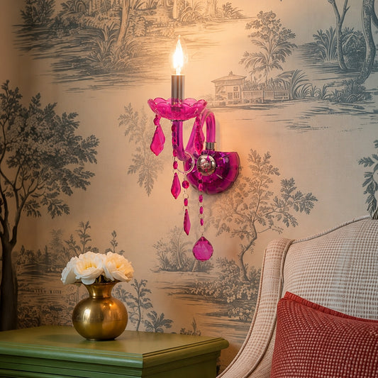 A bright magenta pink acrylic wall sconce with dangling teardrop charms and a lit candle-style bulb, mounted on a beige wallpapered wall above a green side table with a gold vase of white flowers.