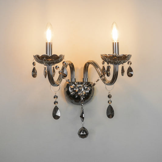A two-light wall sconce with curved silver-toned metal arms, clear faceted crystal teardrop pendants, and lit candle-style bulbs, mounted against a plain light wall.