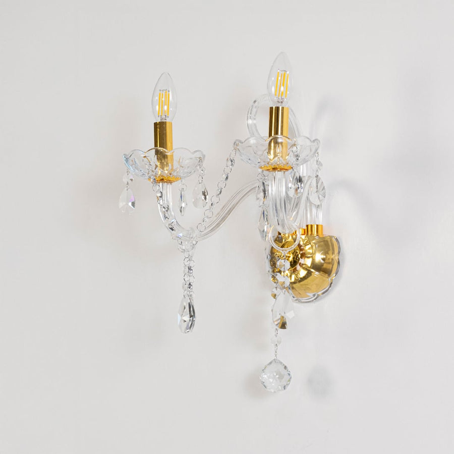 Crystal wall light with golden base dual candle design with crystal drops shown on off white background.