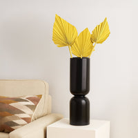 A tall matte black vase displaying three yellow dried palm leaves stands on a white cube table beside a beige sofa with a patterned cushion.