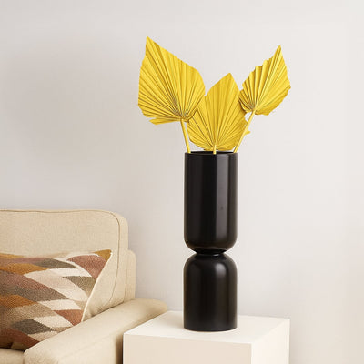 A tall matte black vase displaying three yellow dried palm leaves stands on a white cube table beside a beige sofa with a patterned cushion.