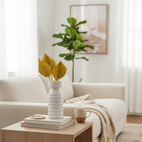 A white ribbed vase holding yellow palm leaves sits on stacked books on a wooden side table in front of a beige sofa and green plant.