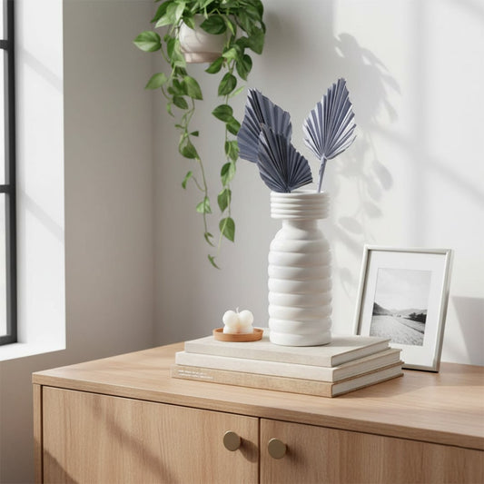 White textured ceramic vase with folded palm leaves styled on a wooden cabinet with books plants and soft daylight in a minimalist home interior.