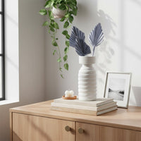 White textured ceramic vase with folded palm leaves styled on a wooden cabinet with books plants and soft daylight in a minimalist home interior.