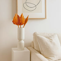 White ceramic vase with orange palm leaves displayed on a pedestal beside a sofa creating a clean minimalist living room decor scene.