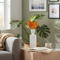 White sculptural ceramic vase with orange palm leaves placed on a wooden side table beside a sofa creating a warm modern living room setting.