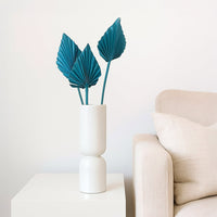 A white ceramic vase displaying three teal palm leaves rests on a white cube table beside a beige armchair with a cushion near a wall.